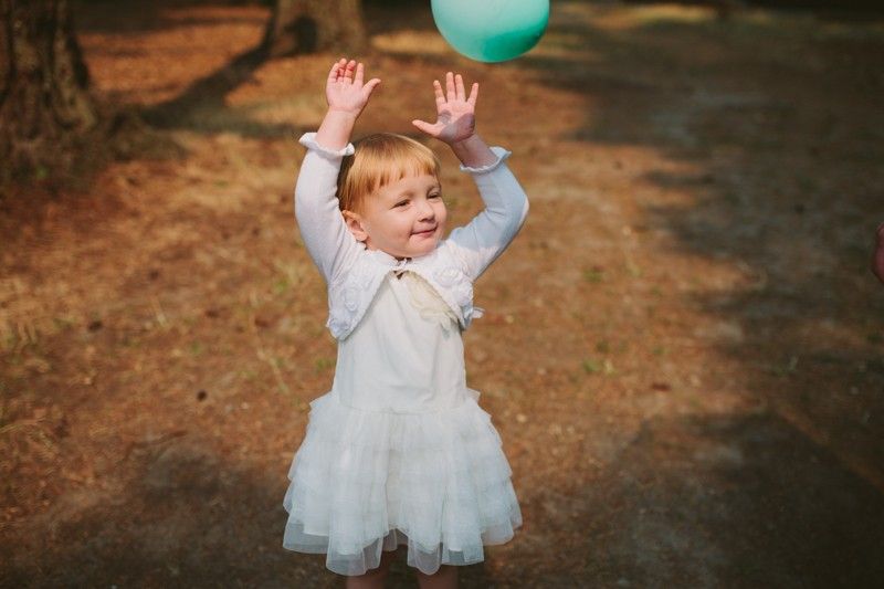 Toddler playing with a balloon at a park wedding, wearing a ruffled white dress and matching cardigan. 