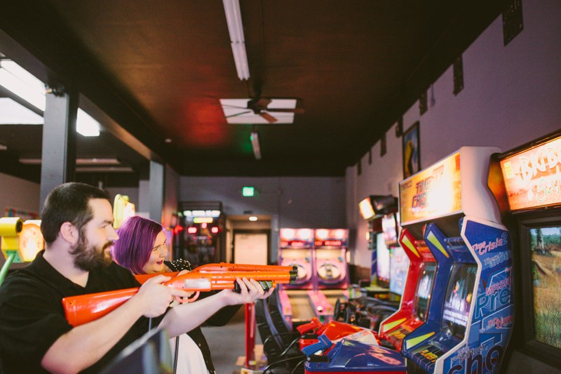 Non-traditional engagement session in an arcade, with bride and groom playing video games. 