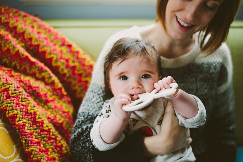Mom holding baby girl, sitting on a colorful couch with a vibrant knit blanket. 