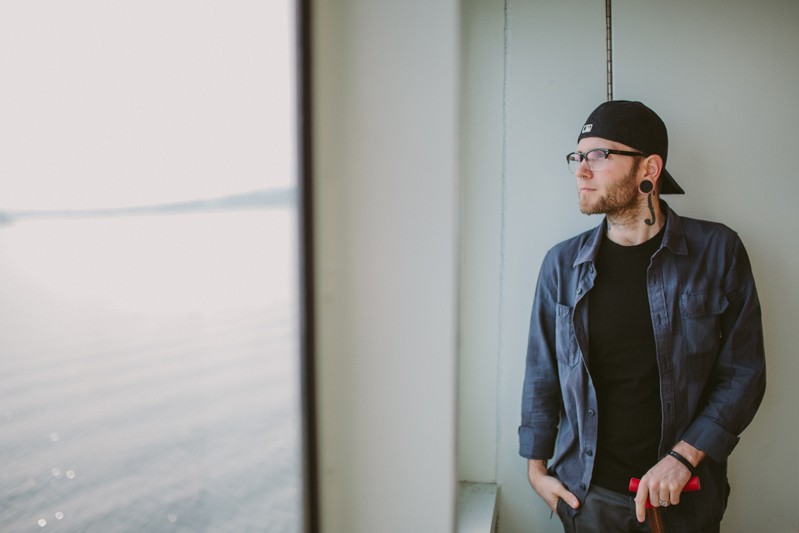 Tattooed man on a ferry, wearing a baseball hat and button up shirt.